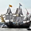 Een groot, historisch zeilschip met drie masten vaart op een rivier of kanaal. De zeilen hangen los en op de masten wapperen Nederlandse en andere vlaggen. In de achtergrond zijn gele hijskranen en windmolens zichtbaar. Er varen meerdere kleine boten vol mensen rondom het schip.