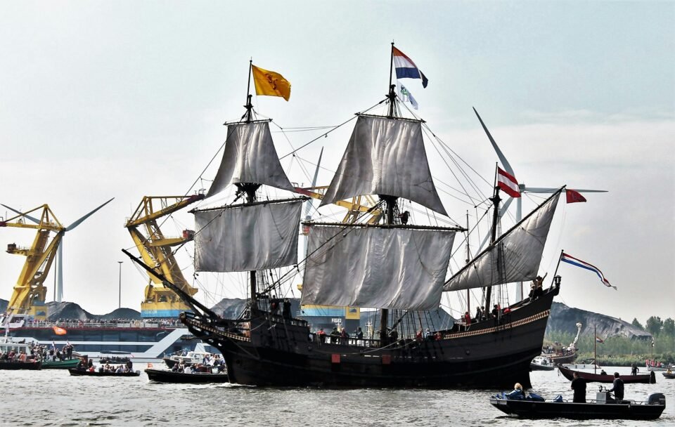 A large, historic sailing ship with three masts is sailing on a river or canal. The sails are hanging loose and Dutch and other flags fly on the masts. Yellow cranes and windmills are visible in the background. Several small boats full of people sail around the ship.