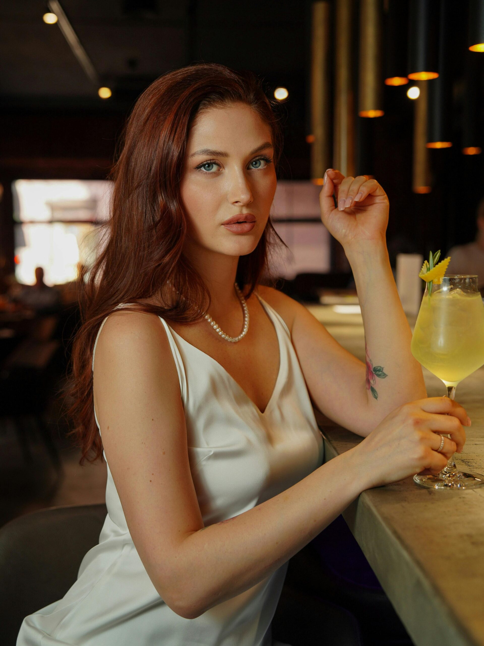 Een vrouw met lang, golvend bruin haar zit aan een bar in een moderne omgeving. Ze draagt een elegante witte top en een parelketting. In haar hand houdt ze een cocktail met een schijfje ananas. De achtergrond is sfeervol verlicht met warme lichtjes.